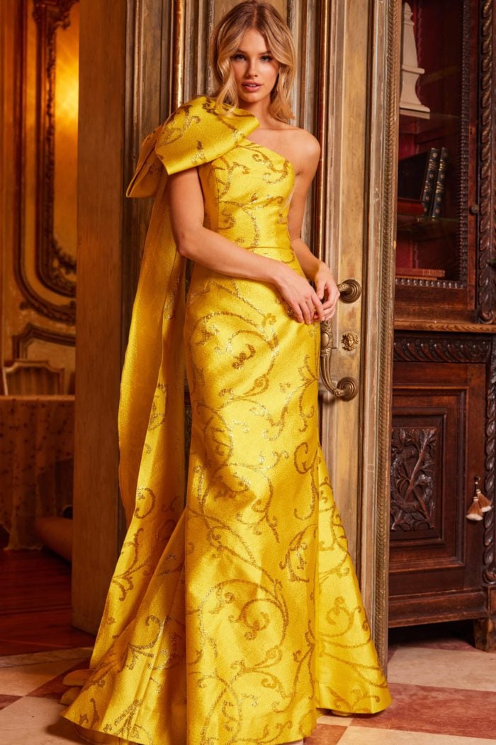 Model wearing Jovani 23742 yellow gown with intricate patterns and bold shoulder design.