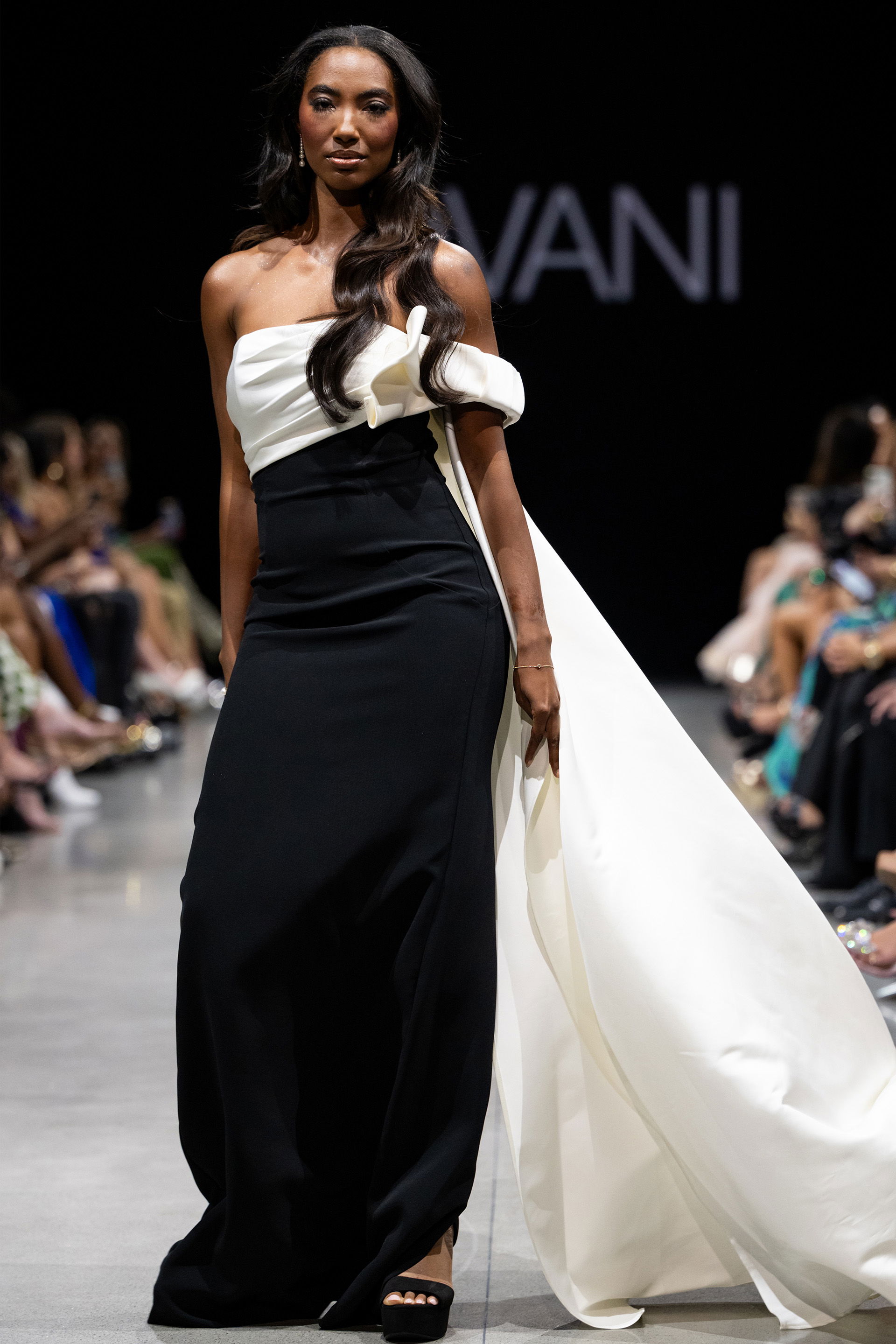 AT38864.jpg Model wearing Jovani AT38864 elegant black gown with asymmetrical neckline and cape-like detail.