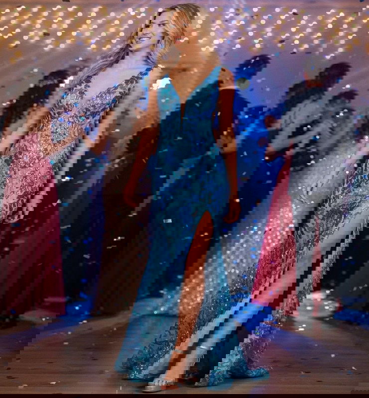Young woman in a sparkly blue prom dress with a high slit, walking on a dance floor with confetti.