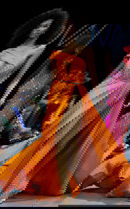 Model wearing S38441 orange gown with off-the-shoulder neckline and high slit